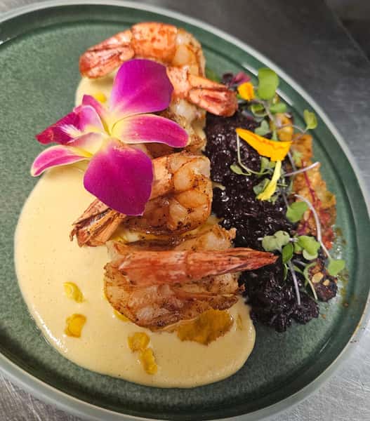 Grilled shrimp served over creamy corn sauce with black forbidden rice, crispy vegetables, microgreens, and a bright pink orchid on a green ceramic plate.