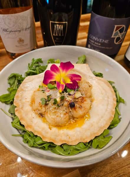 Seared scallops in a shell over a bed of arugula, topped with microgreens and edible flower garnish, served with a curated wine flight in the background.
