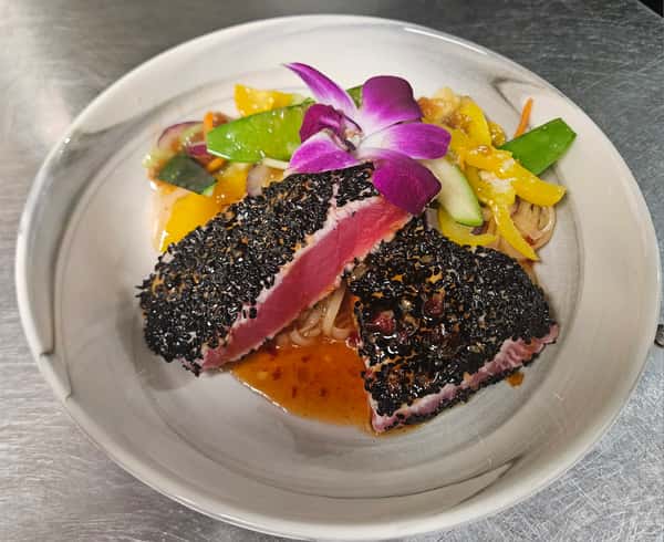 Sesame-crusted ahi tuna served rare over soba noodles with colorful stir-fried vegetables, sweet chili sauce, and a purple orchid garnish on a white bowl plate.