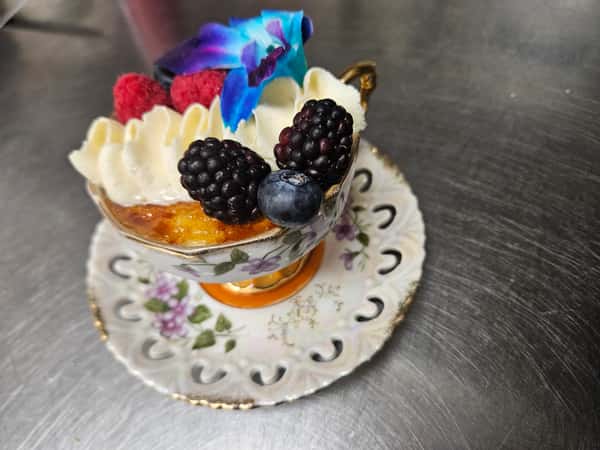 Crème brûlée served in an ornate floral teacup, topped with whipped cream, fresh blackberries, raspberries, a blueberry, and a vibrant blue orchid, placed on a matching vintage saucer.