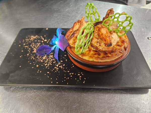 Shrimp-topped gratin in a ramekin with green lattice crisps and a purple orchid, plated on a black rectangular dish with decorative cocoa powder.