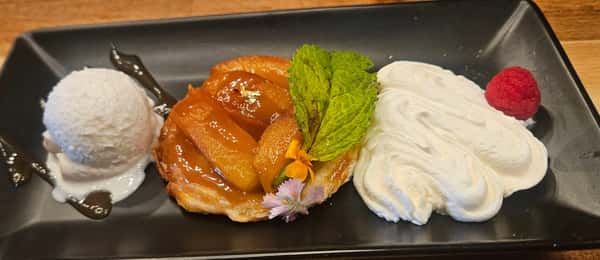 Caramelized apple tart served with whipped cream, vanilla ice cream, a fresh mint sprig, edible flowers, and a raspberry on a sleek black rectangular plate.