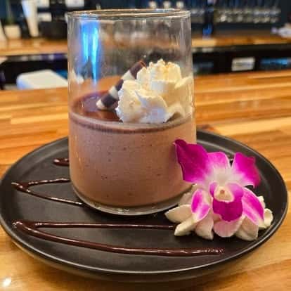 Chocolate mousse dessert in a glass topped with whipped cream and chocolate curls, served on a black plate with chocolate drizzle and a pink orchid flower garnish.