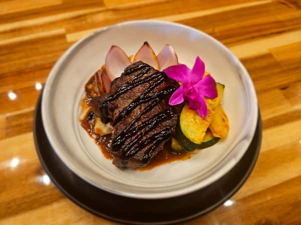 Grilled filet mignon drizzled with balsamic glaze, served with roasted vegetables, caramelized onions, and mashed potatoes, garnished with a purple orchid on a ceramic bowl-style plate.
