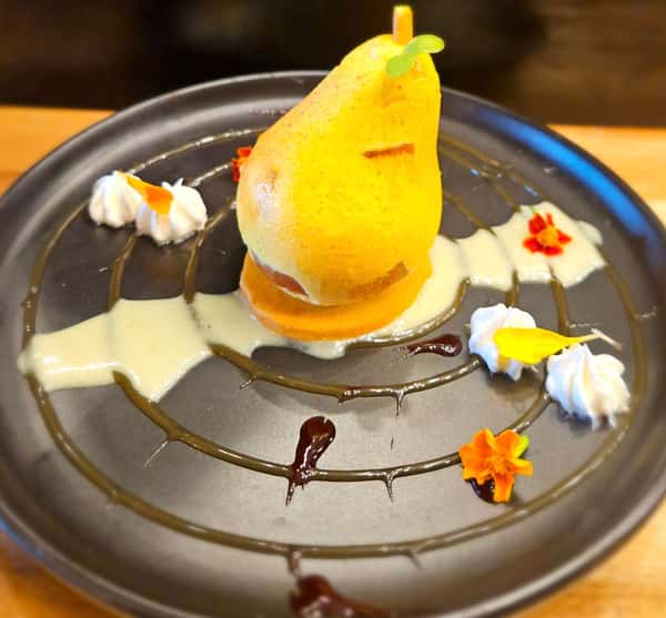 Poached pear dessert plated with whipped cream, edible flowers, and drizzled sauces on a dark round plate in an artistic, modern presentation.