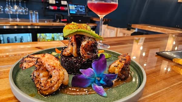Grilled shrimp served over black rice and topped with avocado, sesame seeds, and a vibrant blue orchid, with a glass of rosé wine in the background on a bar counter.