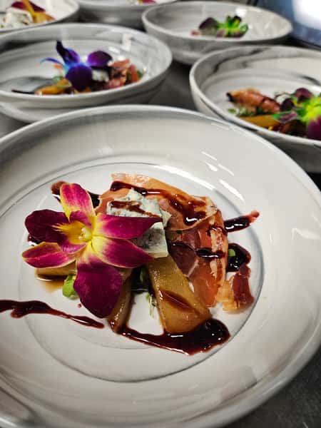 Elegant appetizer featuring prosciutto, poached pear slices, gorgonzola cheese, and balsamic reduction, garnished with an edible orchid flower and plated for service in multiple matching white ceramic bowls.