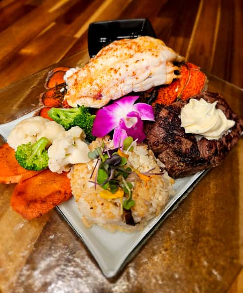 Gourmet surf and turf plate featuring a broiled lobster tail, grilled filet mignon topped with herbed butter, vegetable medley, seasoned rice, and sweet potato slices, garnished with an orchid flower and microgreens, served on a glass plate over a wooden table.