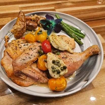 Roasted half chicken served with heirloom tomatoes, artichoke hearts, green beans, crispy potatoes, and capers, garnished with a purple orchid on a ceramic plate.