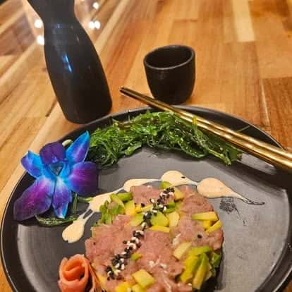 Ahi tuna tartare topped with avocado and sesame seeds, served with seaweed salad, wasabi aioli, soy dipping sauce, and a purple orchid garnish, styled with chopsticks and sake set on a black plate.