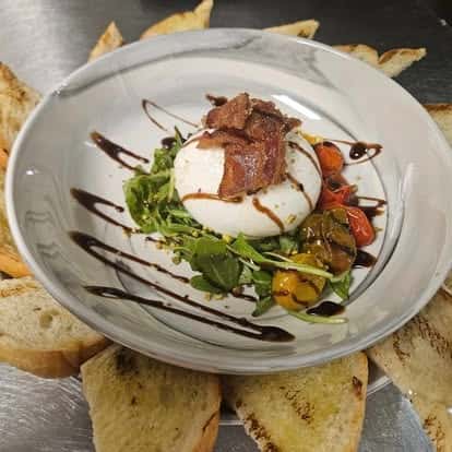 Fresh burrata over arugula and roasted tomatoes, topped with crispy bacon and balsamic glaze, surrounded by grilled crostini on a white ceramic plate.
