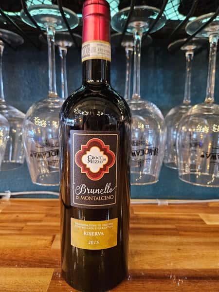 Bottle of Croce di Mezzo Brunello di Montalcino Riserva 2015 wine displayed on a wooden bar with hanging glassware and The Hideaway branding in the background.