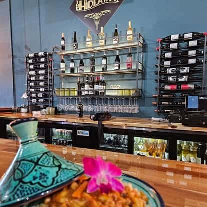 Interior view of The Hideaway wine bar showcasing a sleek bar setup with wine bottles, glassware, and wine racks on a teal-blue wall, with a Moroccan-style tagine dish and orchid in the foreground.