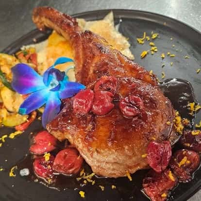 Glazed tomahawk pork chop with cherry compote, served with roasted vegetables and potatoes, garnished with orange zest and a vibrant blue orchid on a black plate.