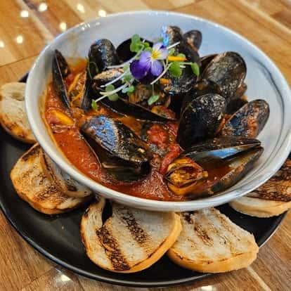 Steamed mussels served in a rich tomato broth with herbs and microgreens, garnished with an edible flower and accompanied by toasted crostini on a black plate over a wooden table.