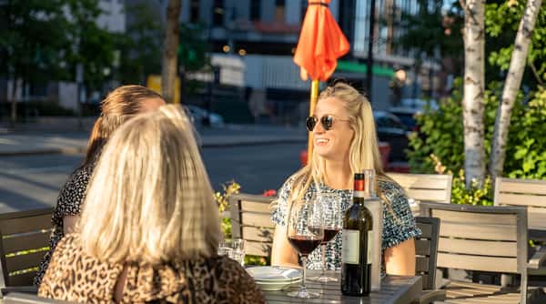 Two women seated at an outdoor restaurant table with drinks in the sunlight.