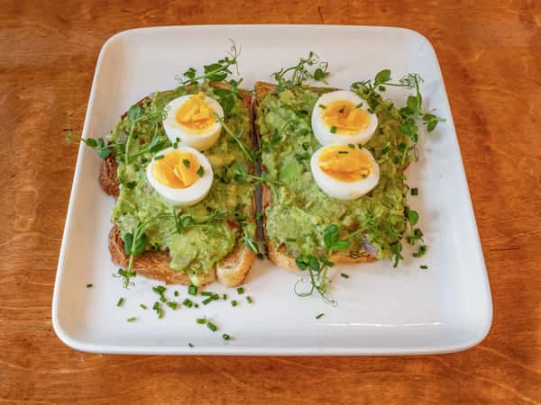 Avocado toast topped with sliced boiled eggs and microgreens on a square plate.