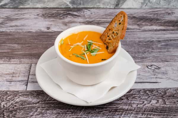 Creamy tomato soup in a white cup, topped with shredded cheese and basil, served with toasted bread slice