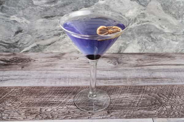 Purple martini in a stemmed glass garnished with a dried citrus slice on a rustic wooden table