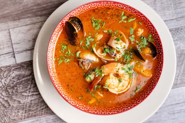 Seafood stew in a red patterned bowl with shrimp, mussels, scallops, and herbs on a white plate