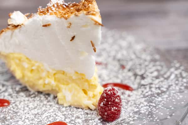 Slice of coconut cream pie topped with whipped cream and toasted coconut, dusted with powdered sugar and raspberries