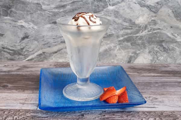 Vanilla ice cream sundae with chocolate drizzle in frosted glass, served with fresh strawberry slices on blue plate