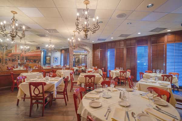 Elegant dining room with white tablecloths, red chairs, chandeliers, and large shuttered windows along brick walls