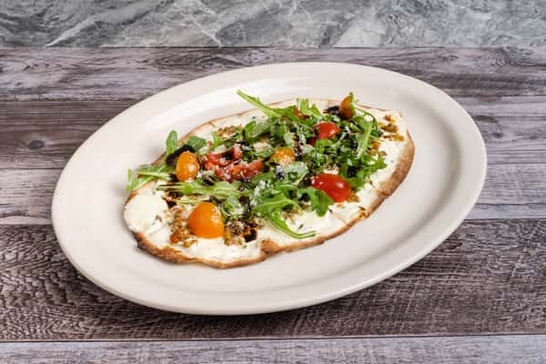 Flatbread pizza topped with arugula, cherry tomatoes, ricotta, and balsamic glaze on white plate