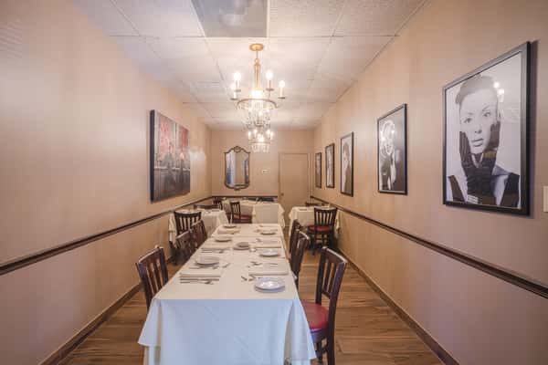 Narrow private dining room with white tablecloth tables, chandeliers, wood floors, and framed portraits on beige walls