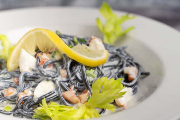 Black squid ink pasta in creamy sauce with seafood, topped with lemon slices and celery leaves