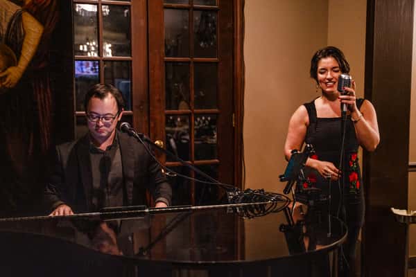 Male pianist playing a grand piano beside female singer holding vintage microphone in cozy restaurant setting