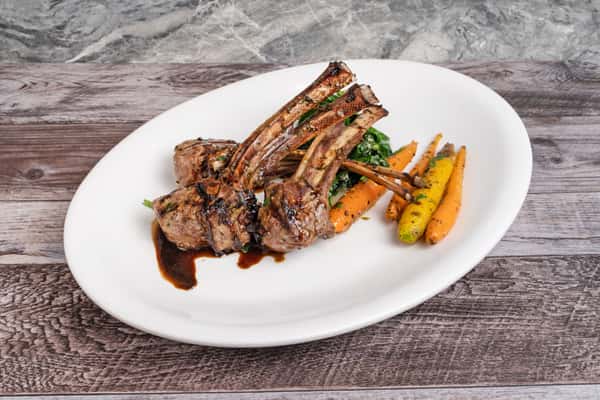 Grilled lamb chops with rosemary jus, served with sautéed greens and roasted rainbow carrots on white plate