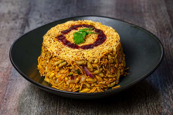 Indian street-style bhel puri snack made with puffed rice, sev, chutney swirl, onions, and cilantro on a black plate.