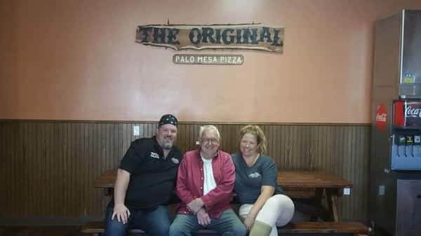 Three people smiling on a bench inside a rustic pizza restaurant beneath a "The Original Palo Mesa Pizza" sign, with wood panel walls and a soda fountain machine nearby.