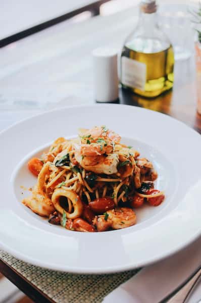 Seafood pasta with shrimp, calamari, and cherry tomatoes, garnished with herbs, served in a white bowl on a wooden table.