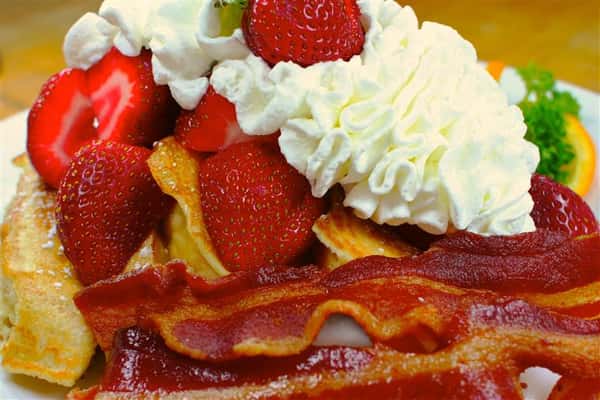 Pancakes topped with strawberries and whipped cream with a side of bacon
