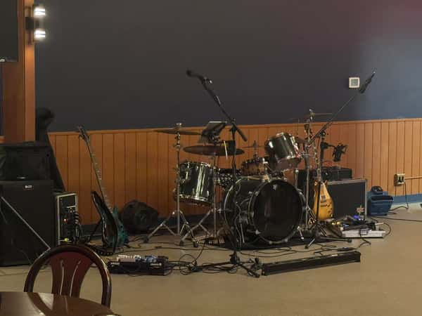 Live music setup with drum kit, guitars, mics, and amplifiers against a dark blue and wood-paneled wall inside a venue.