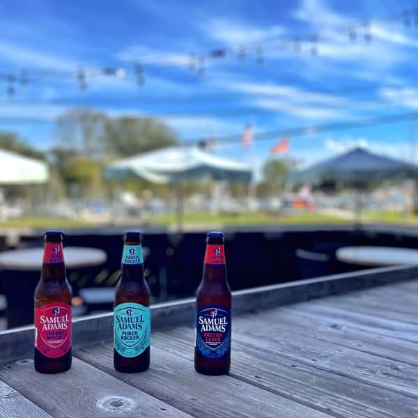 Three Samuel Adams beers on a wooden patio table with string lights and umbrellas in the background on a sunny day.