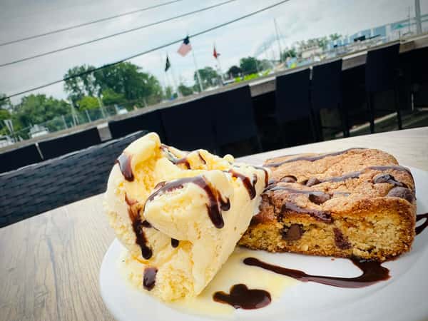 Vanilla ice cream topped with chocolate sauce, served beside a warm chocolate chip cookie bar on a white plate.