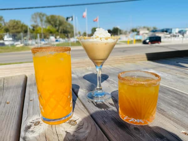 Three colorful cocktails with sugar-rimmed glasses on a wooden table, set outdoors on a sunny day near a marina.