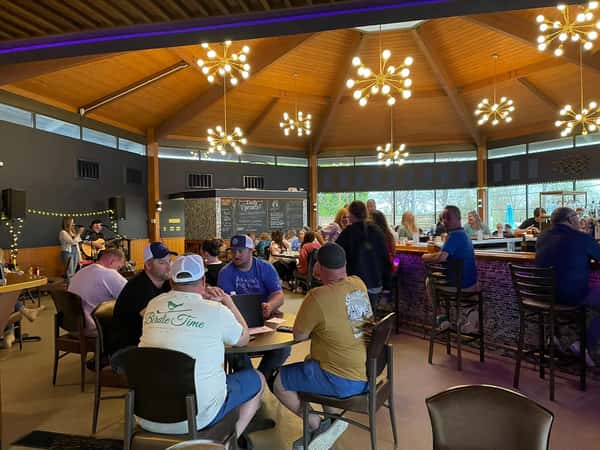 Guests enjoying drinks, food, and live acoustic music inside a warmly lit bar and restaurant with modern chandeliers.