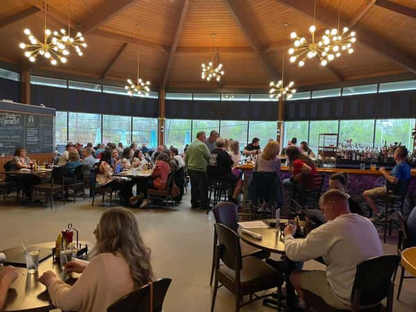 Busy restaurant interior with modern lighting, a central bar, large windows, and patrons dining and socializing at tables.