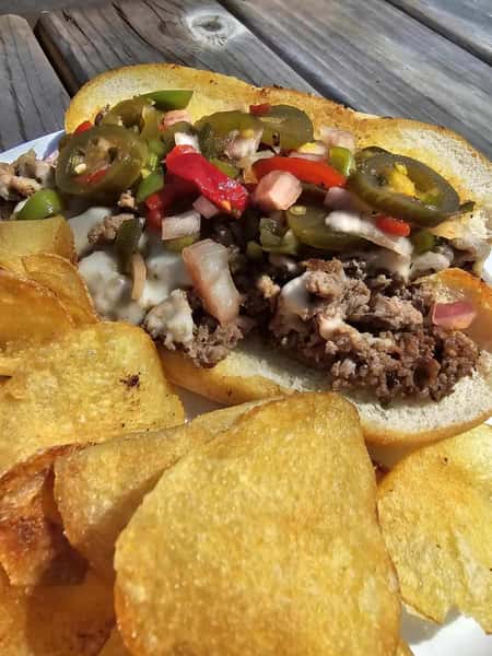 This image features a hearty sandwich filled with seasoned ground beef or steak, melted cheese, sautéed onions and bell peppers, and topped with pickled jalapeños and what appears to be a fresh salsa.