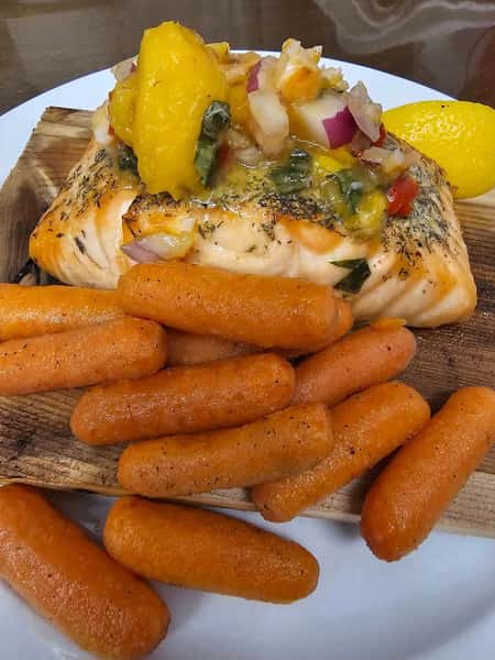 Grilled salmon topped with peach salsa, served with glazed baby carrots and a lemon wedge on a wooden plank.