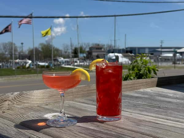 Two red cocktails with lemon garnishes sit on a wooden patio table with a marina and flags in the background.