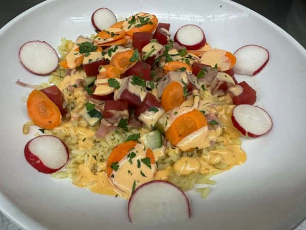 This dish appears to be a poke bowl featuring diced raw tuna, sliced radishes and carrots, cucumber, and a creamy spicy mayo-style dressing over a bed of rice, garnished with chopped herbs.