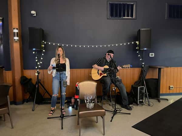 Female vocalist and male guitarist perform live in a cozy indoor setting with string lights and sound equipment.