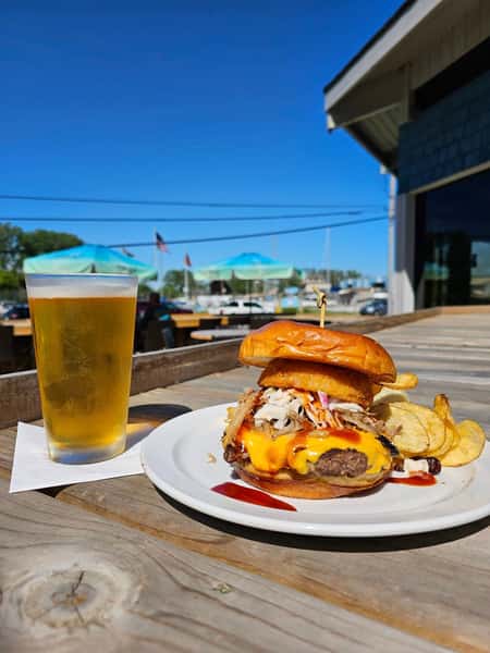 Cheeseburger stacked with pulled pork, coleslaw, and onion ring, served with chips and a pint of beer on a sunny patio.