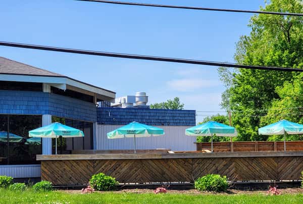 Outdoor patio with turquoise umbrellas, wood railing, and surrounding greenery next to a blue-trimmed restaurant building.