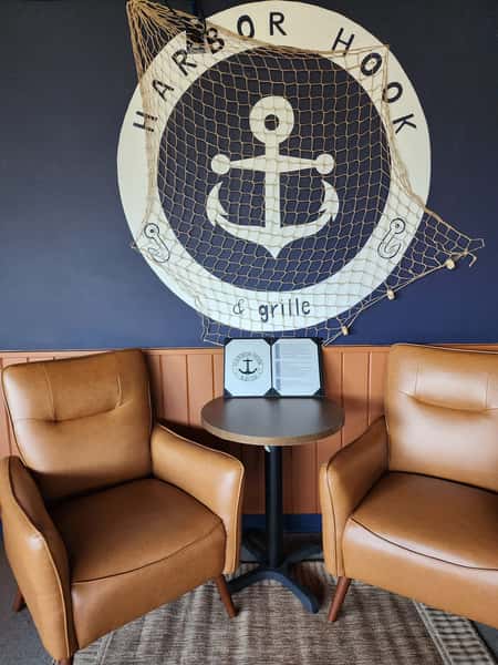 Two brown leather chairs and a table beneath a nautical anchor mural reading "Harbor Hook & Grille" with netting decor.